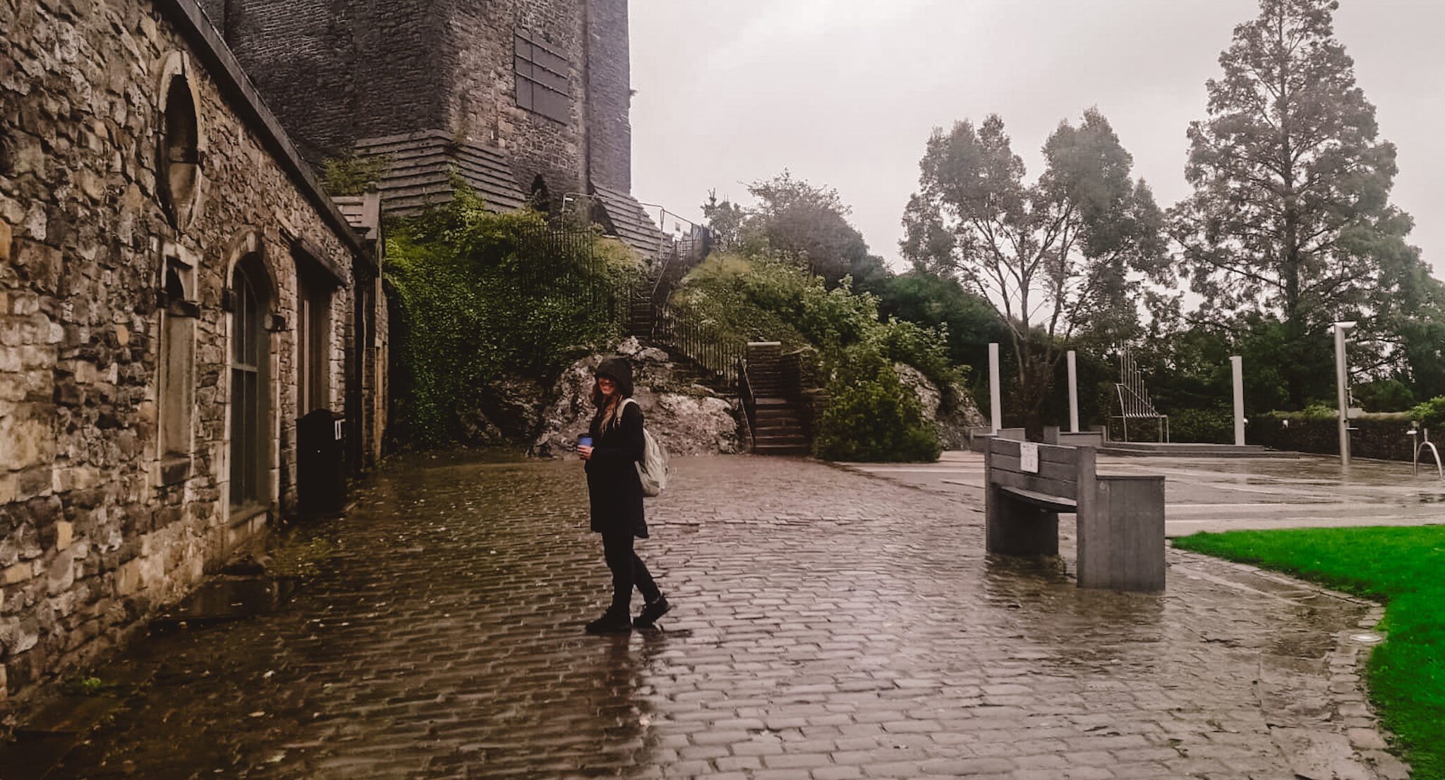 A woman holding a coffee walks across rain-soaked cobblestones beside Clitheroe Castle in Lancashire, England.