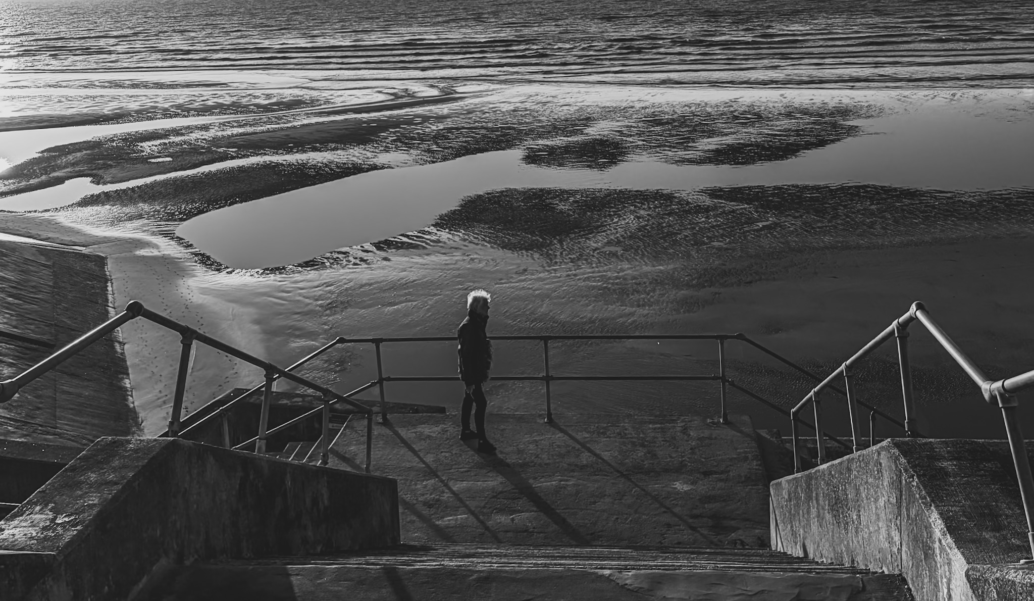 A lone figure standing on a quiet beach on the English coast beneath a wide, overcast sky.