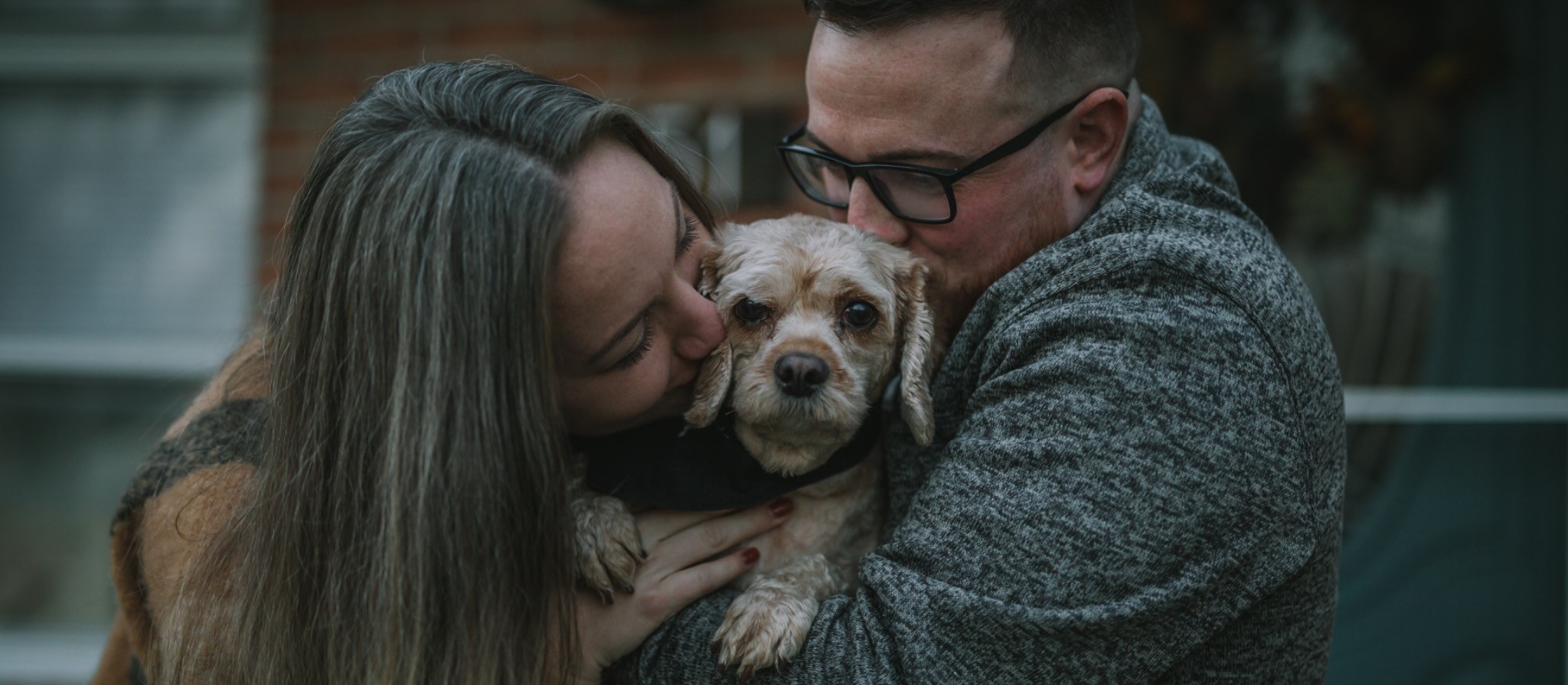 A couple kisses their small dog from either side while holding them close during an outdoor portrait.