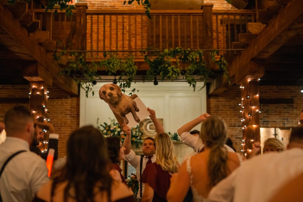 A life-size cutout of a small dog stands on the dance floor among wedding guests during the reception.