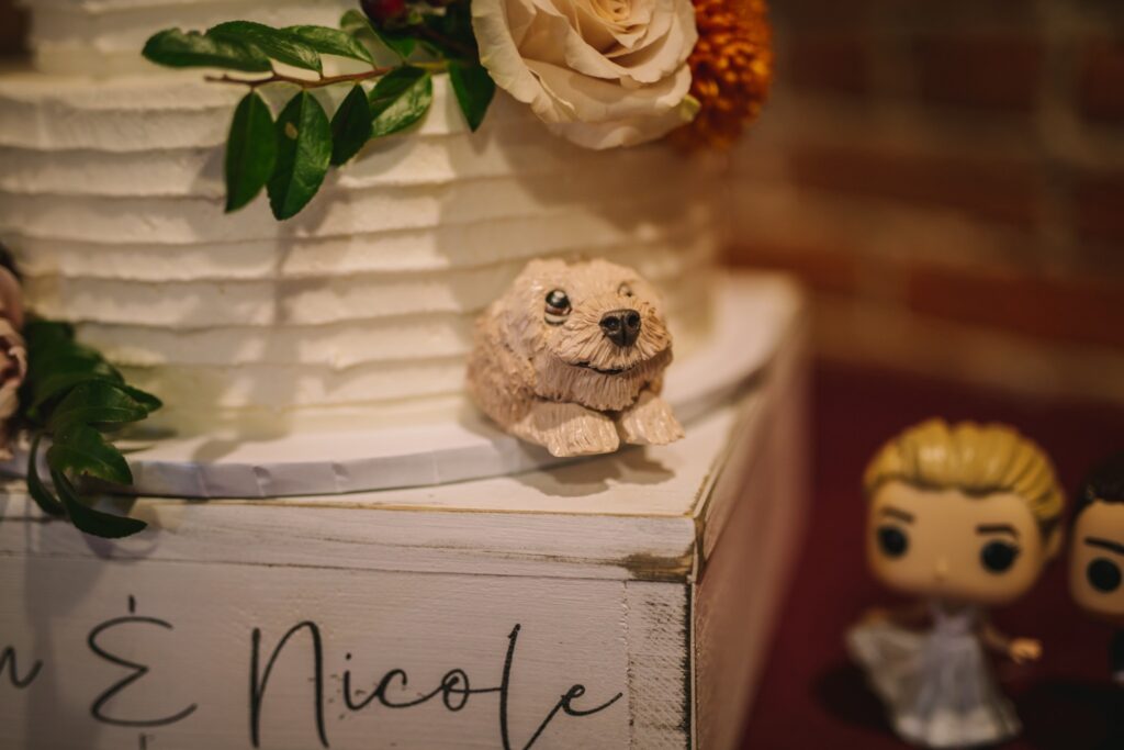 A small dog figurine sits beside a white wedding cake with floral decorations and miniature bride-and-groom figures.